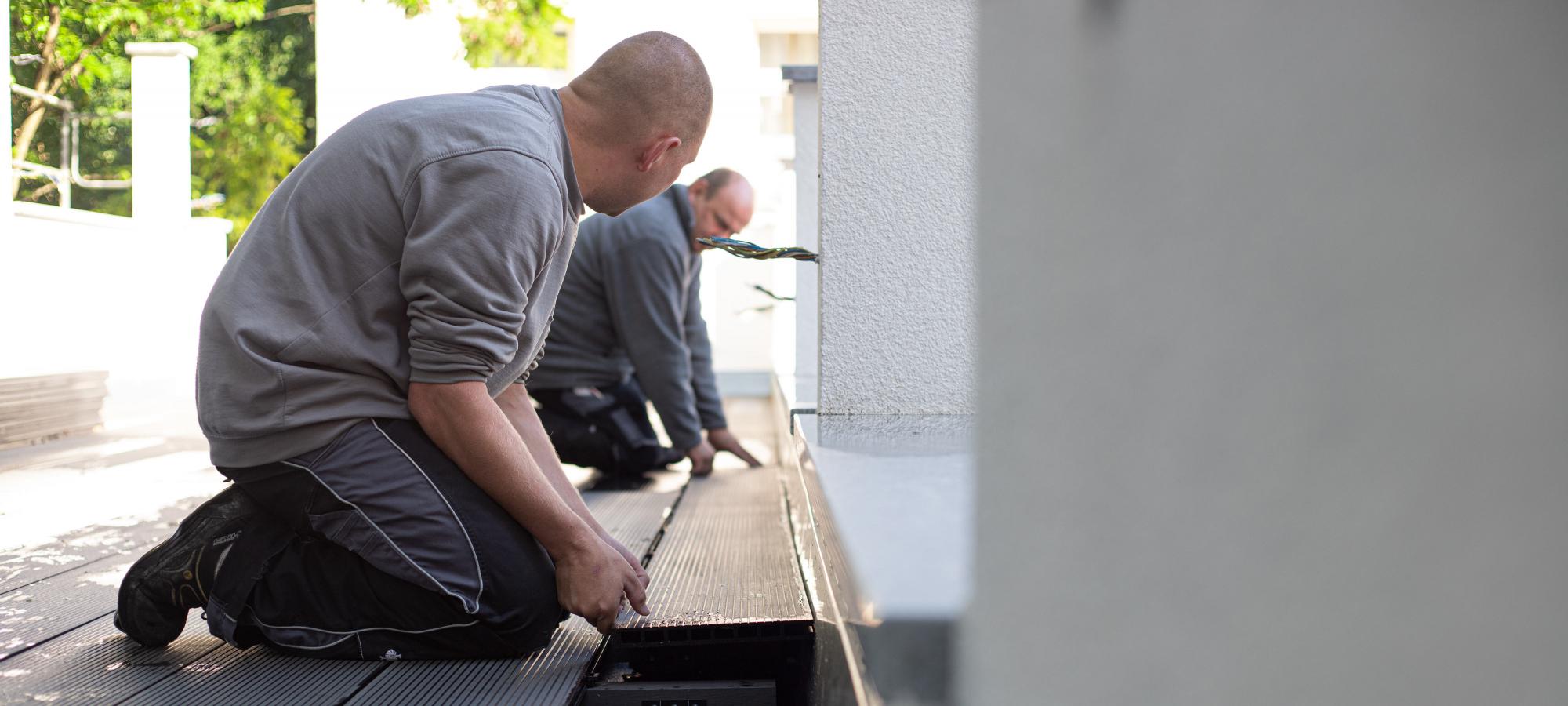Zwei Männer in langer Arbeitskleidung verlegen an einem sonnigen Tag graue WPC-Dielen auf einer Terrasse.
